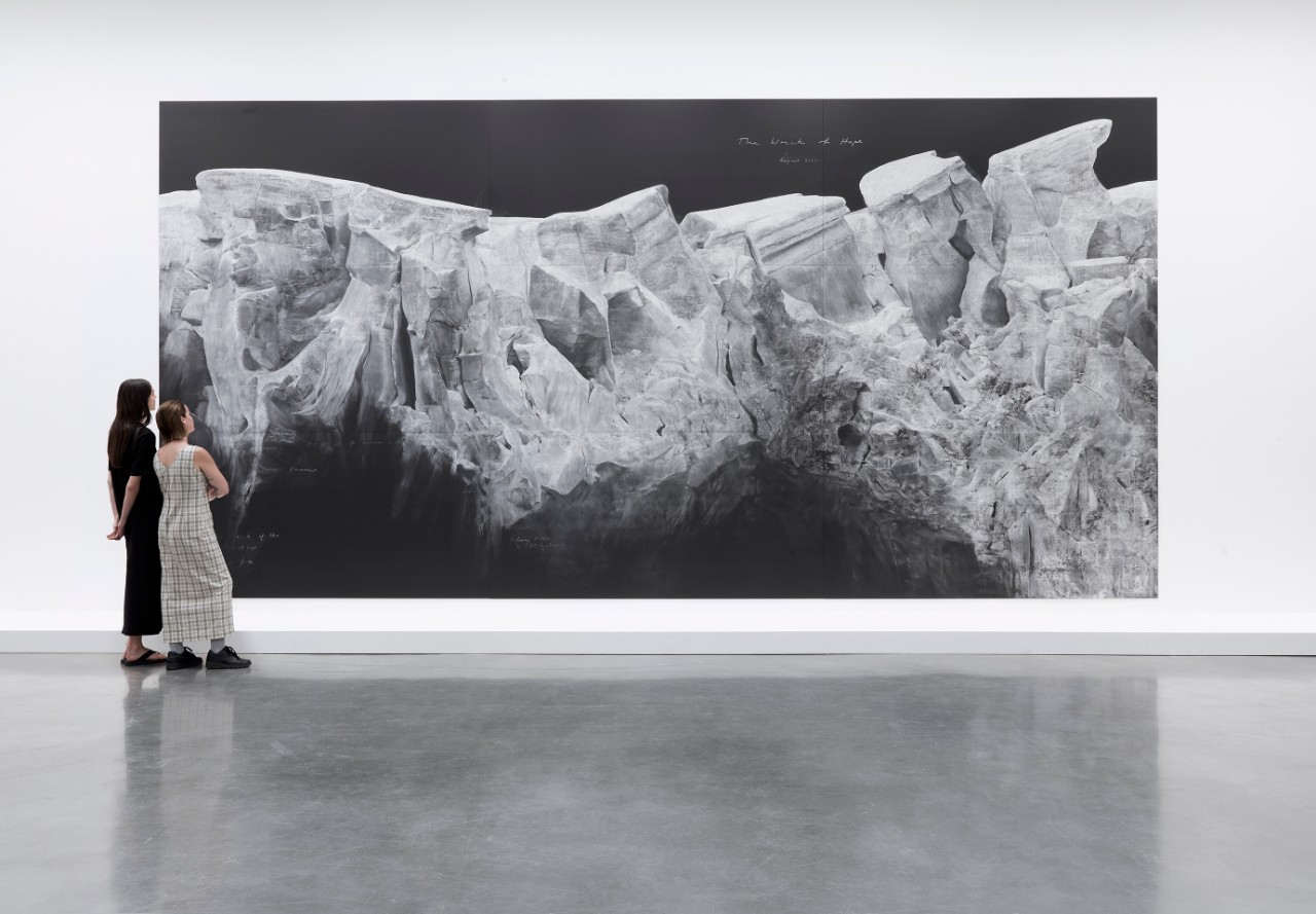 two young women standing in an art gallery looking at a picture on the wall, it looks like a black and white photo of icebergs
