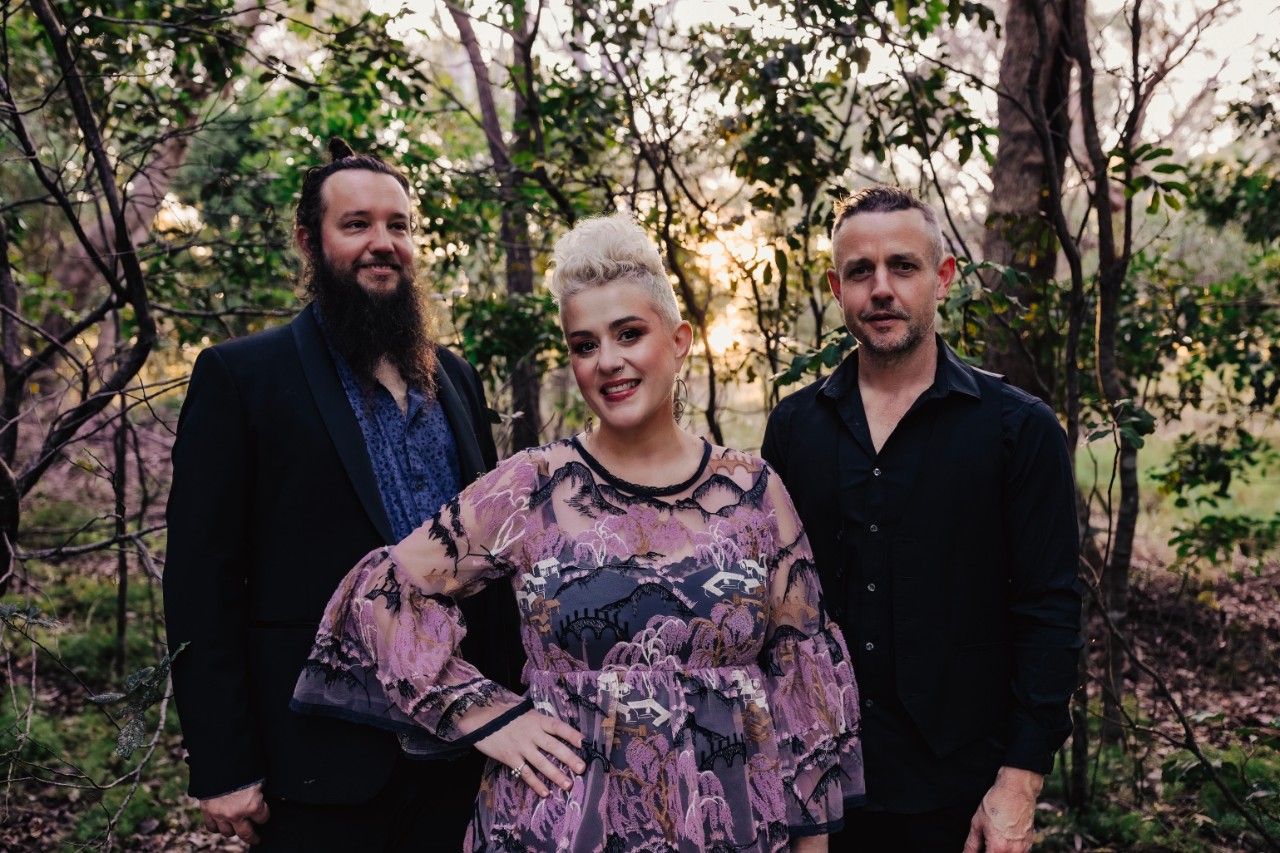 three musicians standing in a garden smiling at the camera