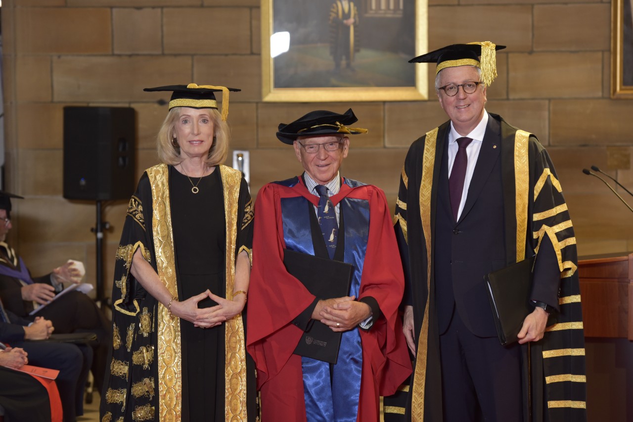 Three people – a woman and two men – dressed in academic robes and standing in front of a standstone wall.