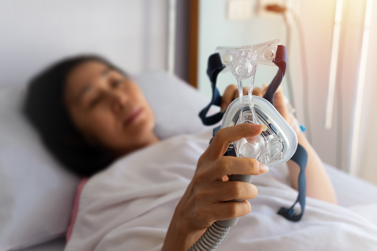 A woman lies in bed holding a CPAP facemask for sleep apnoea therapy